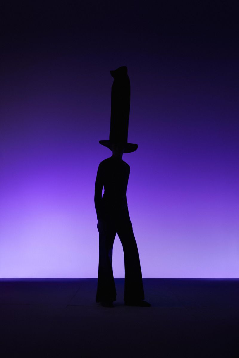 Silhouette of a person with a very tall hat against a gradient purple background, creating a fashion-forward and theatrical image.