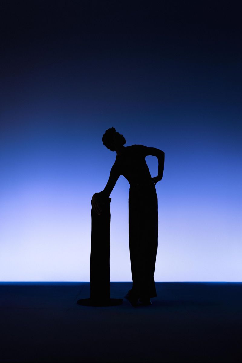 Silhouette of an adult female in long dress leaning on cylindrical object against a gradient blue and white background in a studio.