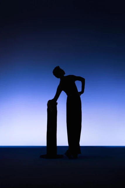 Silhouette of an adult female in long dress leaning on cylindrical object against a gradient blue and white background in a studio.