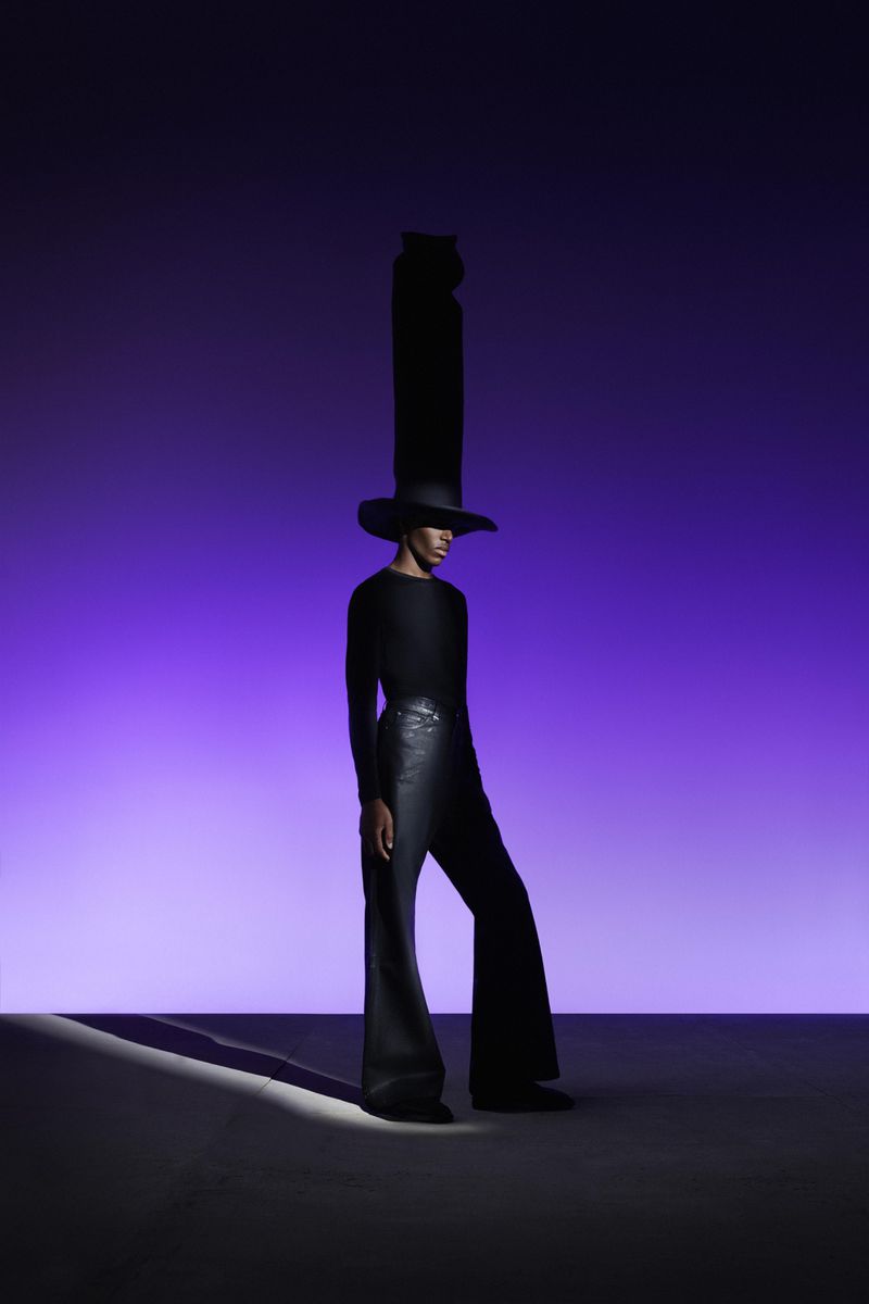 Fashionable person with tall black hat and flared pants poses against a vivid purple gradient backdrop in a studio lighting setting.