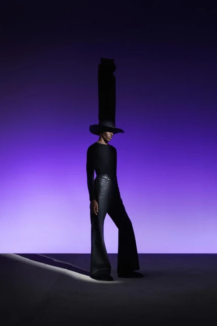 Fashionable person with tall black hat and flared pants poses against a vivid purple gradient backdrop in a studio lighting setting.