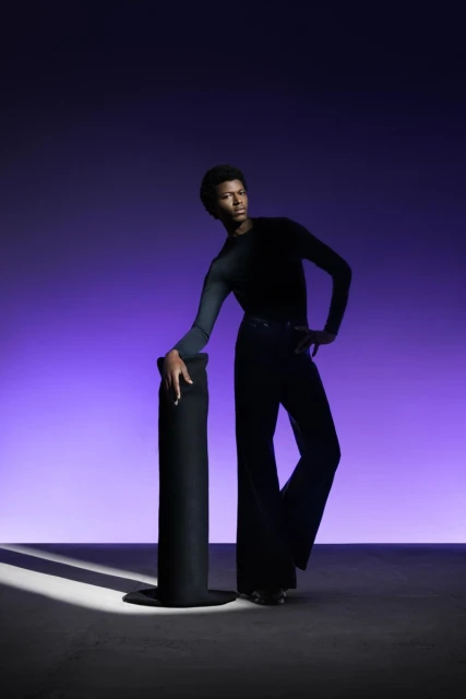 Fashion portrait of an adult with dark skin in a dark top and bell-bottom pants, leaning on a black top hat under studio lighting.