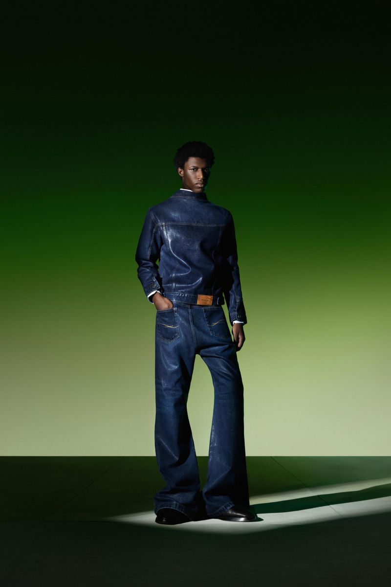 Man in a navy-blue denim outfit stands in a photo studio with green gradient backdrop and a spotlight accentuating the look and fabric.