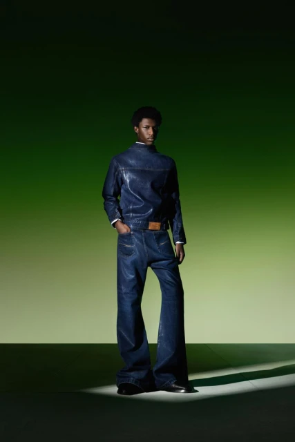 Man in a navy-blue denim outfit stands in a photo studio with green gradient backdrop and a spotlight accentuating the look and fabric.