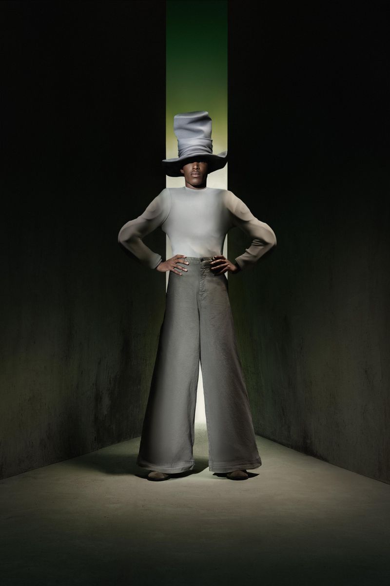 Person wearing a wide-brimmed hat and bell-bottom pants poses in front of a green light in a dark, stylized studio fashion portrait.