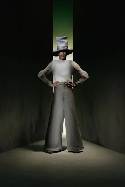 Person wearing a wide-brimmed hat and bell-bottom pants poses in front of a green light in a dark, stylized studio fashion portrait.