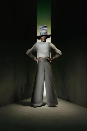 Person wearing a wide-brimmed hat and bell-bottom pants poses in front of a green light in a dark, stylized studio fashion portrait.