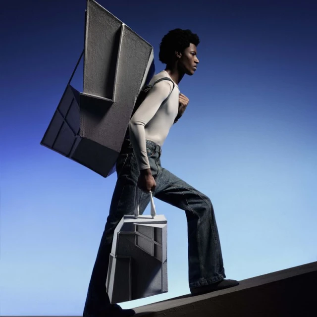Man carries geometric structural cases against blue sky. He wears a white top, jeans, and black shoes. One case is a backpack.