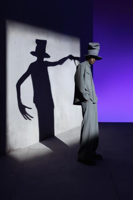 Man in a grey suit and tall hat standing next to a wall with his long-fingered shadow projected on it in a studio setting.