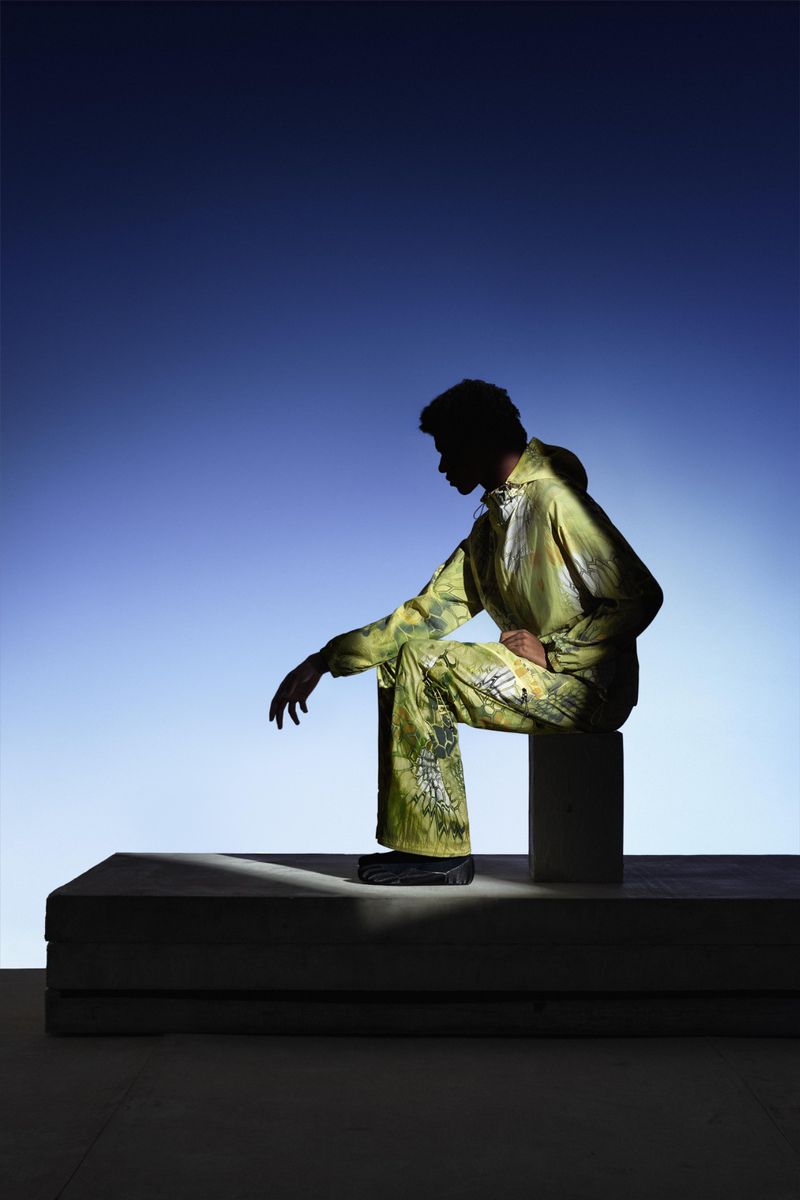 A person is silhouetted against a blue background, wearing a green patterned jacket and pants, and sitting on a white block.