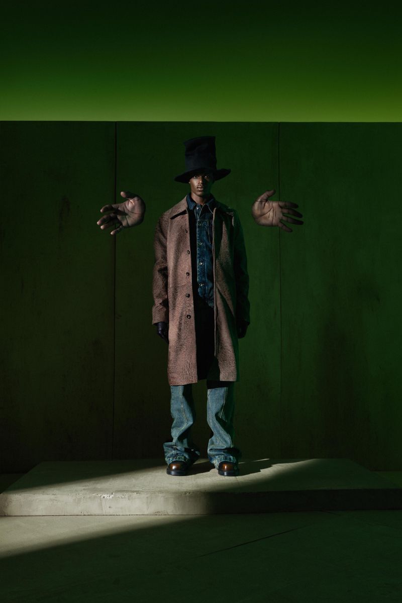 Man in a brown coat, blue jeans, and black top hat stands in front of a green wall with floating hands for a fashion editorial.