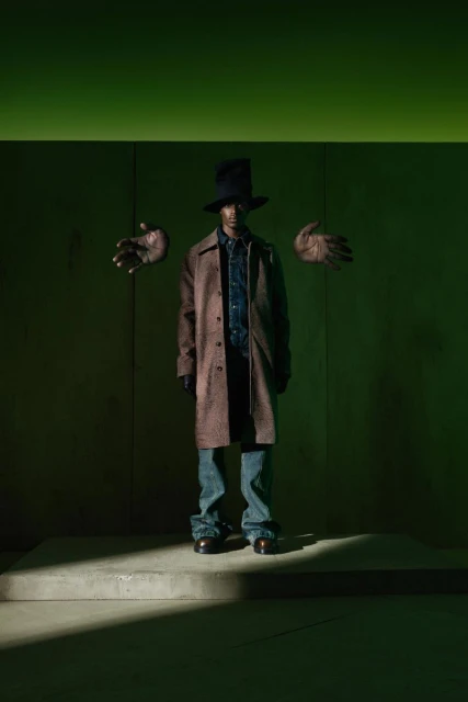 Man in a brown coat, blue jeans, and black top hat stands in front of a green wall with floating hands for a fashion editorial.