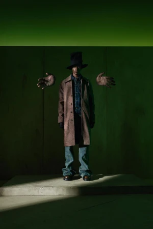 Man in a brown coat, blue jeans, and black top hat stands in front of a green wall with floating hands for a fashion editorial.