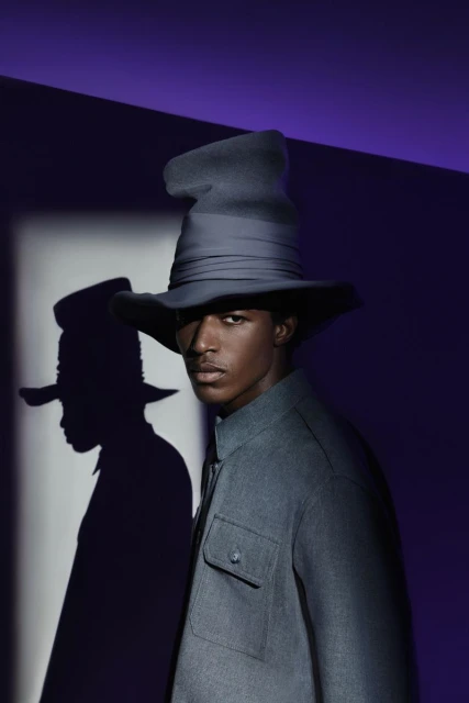 Portrait of a black male fashion model wearing a large hat and gray jacket, standing in a studio with purple and white lighting.