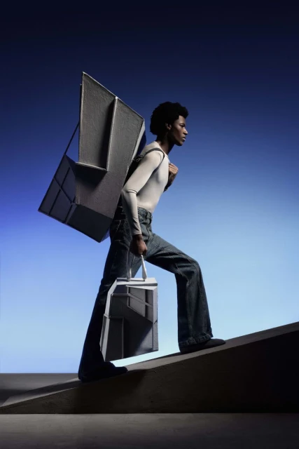 A woman climbs stairs while carrying geometric shapes on her back and in hand, with a gradient blue background and studio lighting.