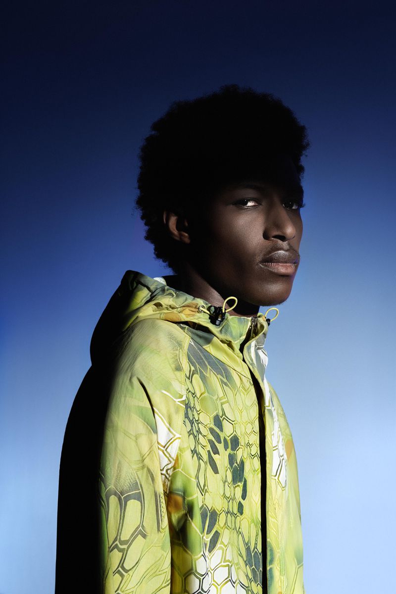 Headshot of an African American man with afro hair, wearing a patterned hooded jacket against a gradient blue backdrop.