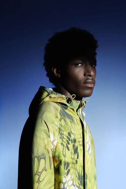 Headshot of an African American man with afro hair, wearing a patterned hooded jacket against a gradient blue backdrop.