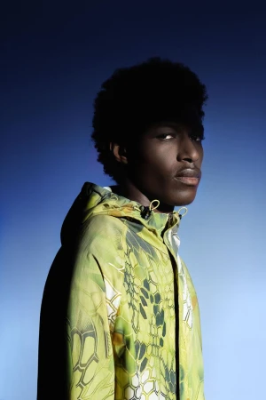 Headshot of an African American man with afro hair, wearing a patterned hooded jacket against a gradient blue backdrop.