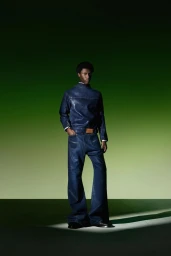 Black man wearing a matching blue denim top and wide-leg trousers standing against a green gradient studio background.