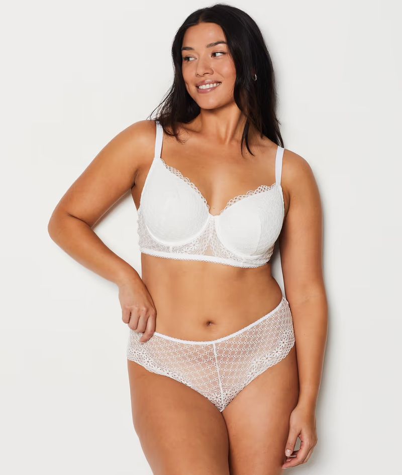 Smiling woman with dark hair poses in a white lace bra and matching underwear against a white background in a studio setting.