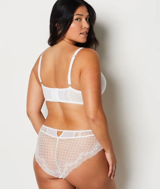 Woman with dark hair poses from behind in a white, lacy bra and matching underwear against a plain white studio backdrop.