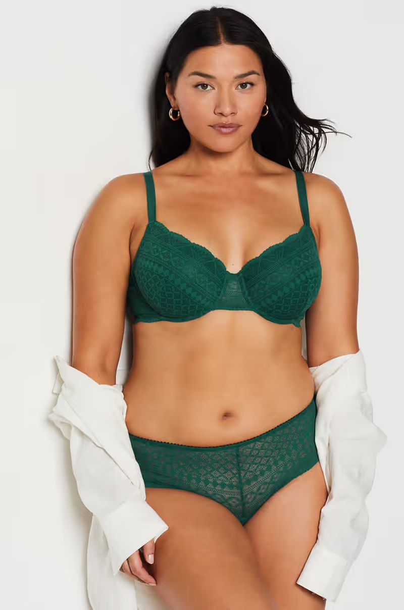 Woman in a green lace bra and panty set poses in a studio against a white wall with a white shirt draped off one shoulder.