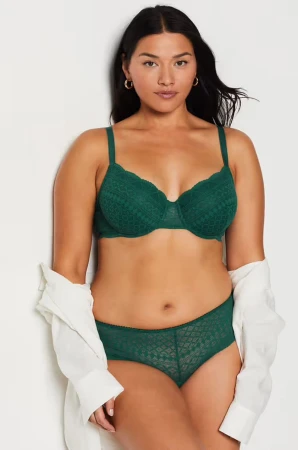 Woman in a green lace bra and panty set poses in a studio against a white wall with a white shirt draped off one shoulder.