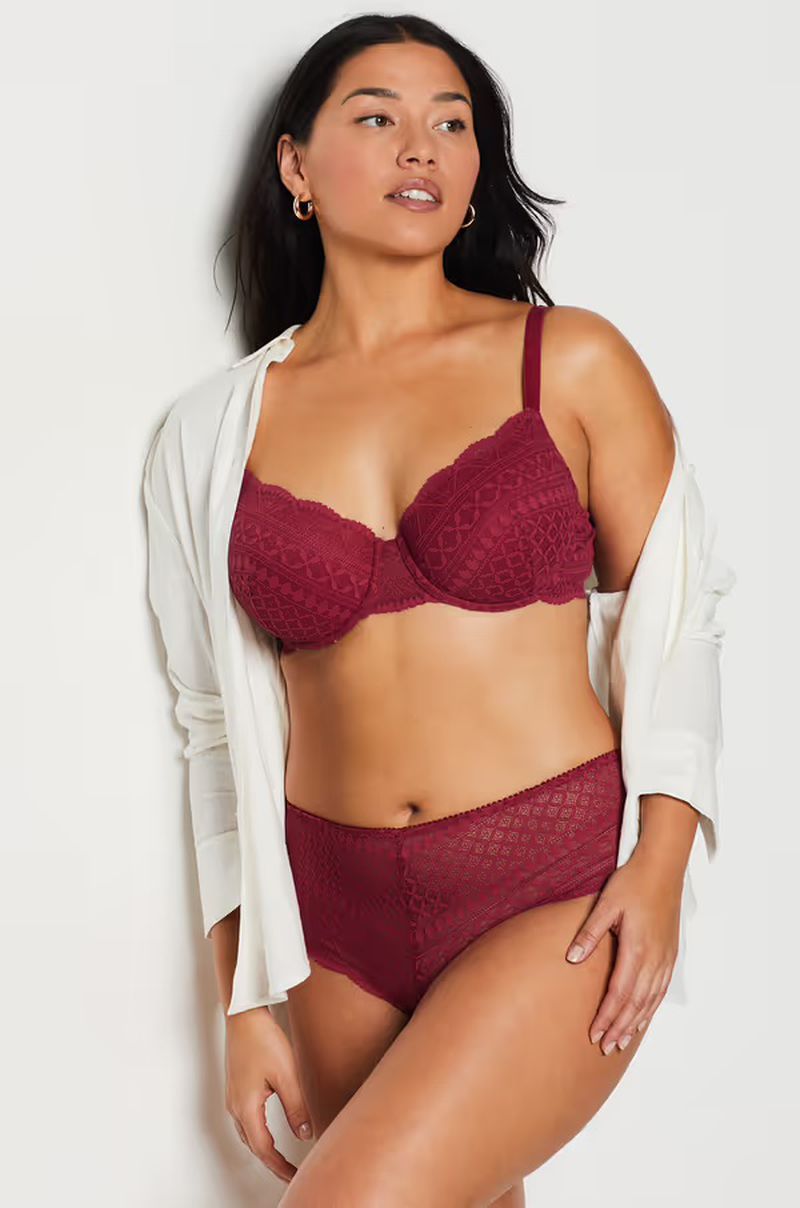 Brunette woman in a red lace bra and underwear set with a white shirt. She poses against a white wall indoors.