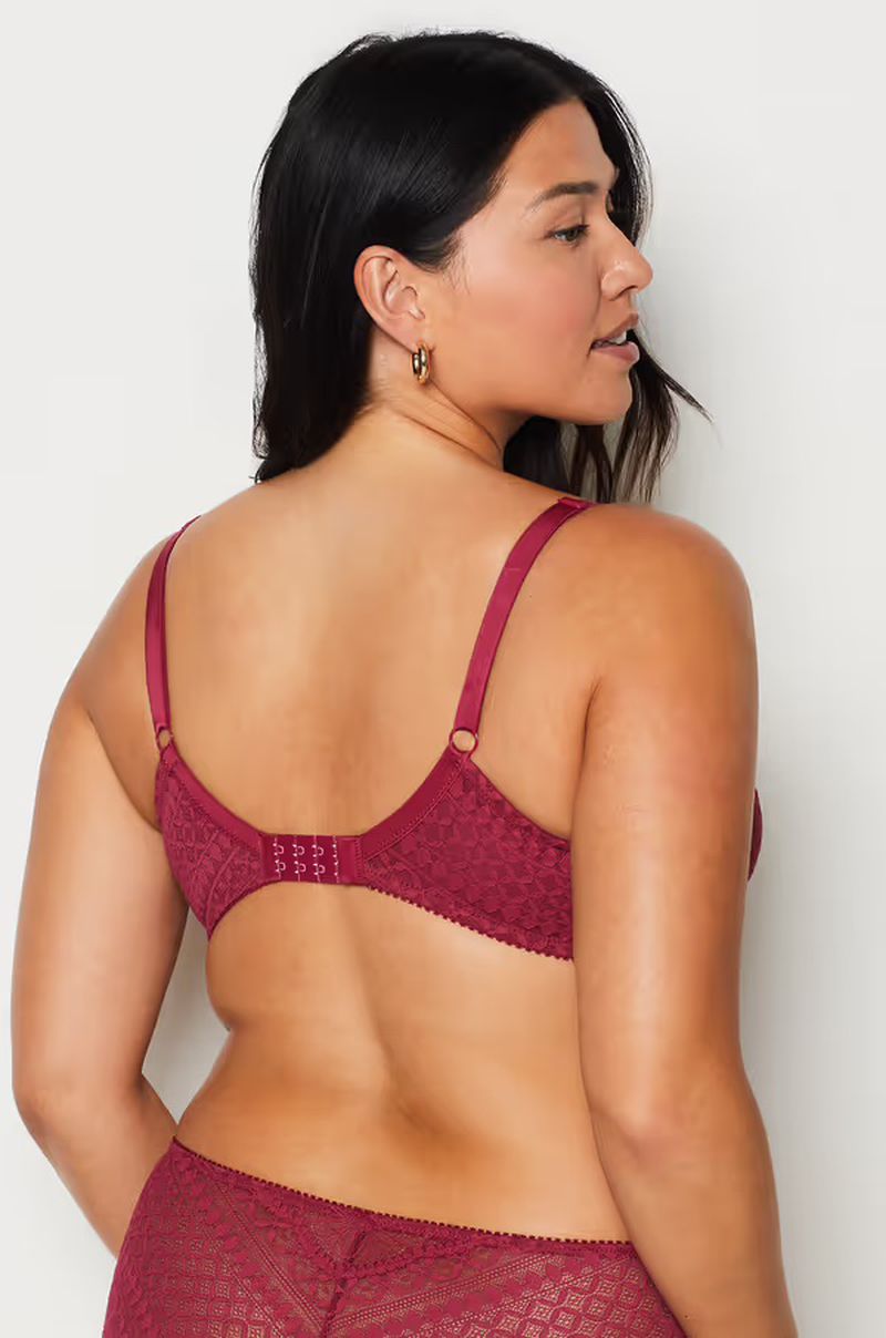 Brunette woman with back to the camera poses in a burgundy-colored lace bra and matching underwear against a plain white backdrop.