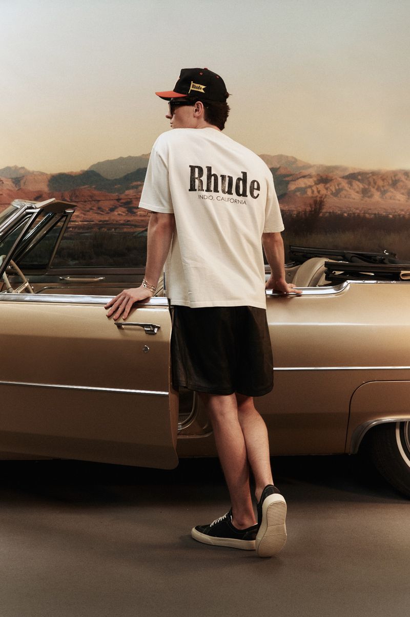 An adult male, viewed from the rear, leans against a classic convertible car in a desert landscape wearing a white t-shirt and baseball cap.