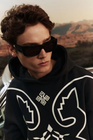 A young man with wavy brown hair poses in a navy hoodie with white graphics and black sunglasses against an out-of-focus outdoor backdrop.