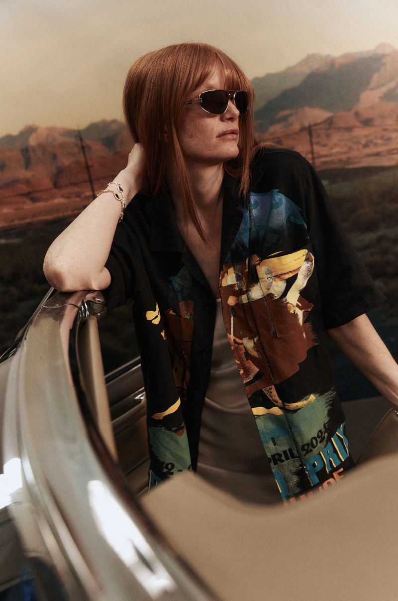 A stylish woman with red hair and sunglasses leans against a boat with a desert and mountain landscape background, showcasing fashion.