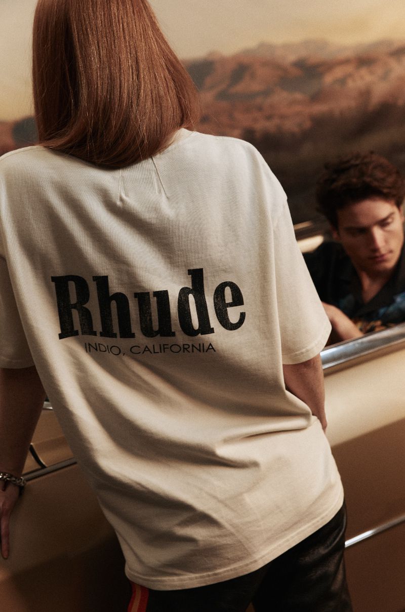 Person in Rhude shirt with desert backdrop; another person is seated nearby. Focus on the shirt and backdrop.
