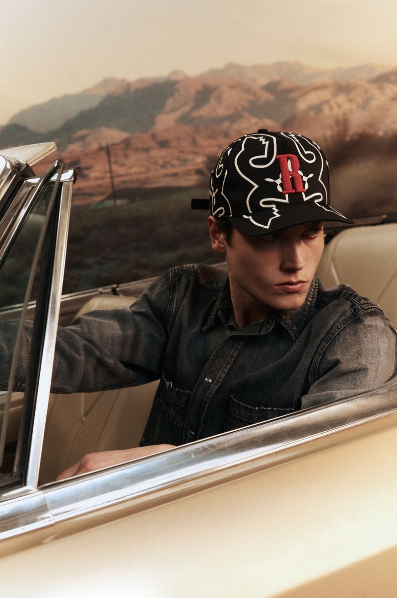 Young man in car wearing patterned baseball cap with letter 'R' in a desert landscape. Fashion photography, vintage automobile.