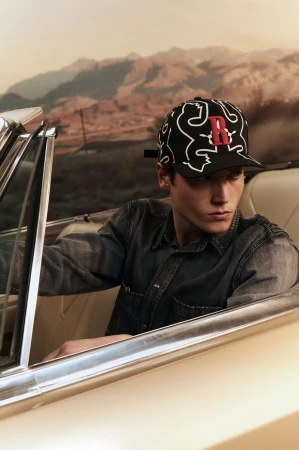 Young man in car wearing patterned baseball cap with letter 'R' in a desert landscape. Fashion photography, vintage automobile.