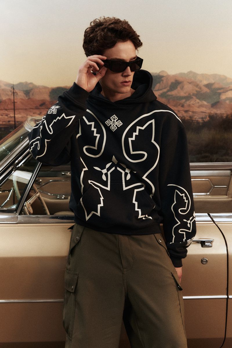 Male model in sunglasses and hoodie leaning against a classic car against a desert background. Fashion editorial photoshoot.