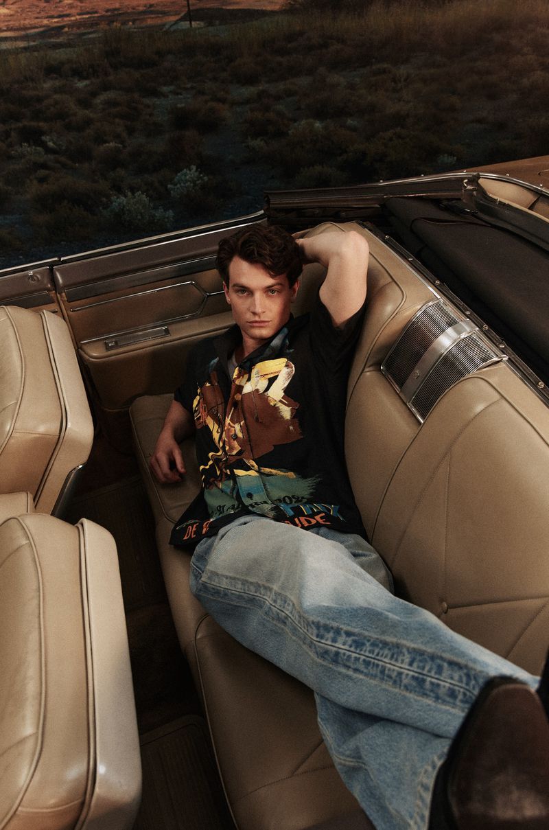 A relaxed adult male is reclined inside a vintage convertible with a dark natural landscape visible in the background during daytime.