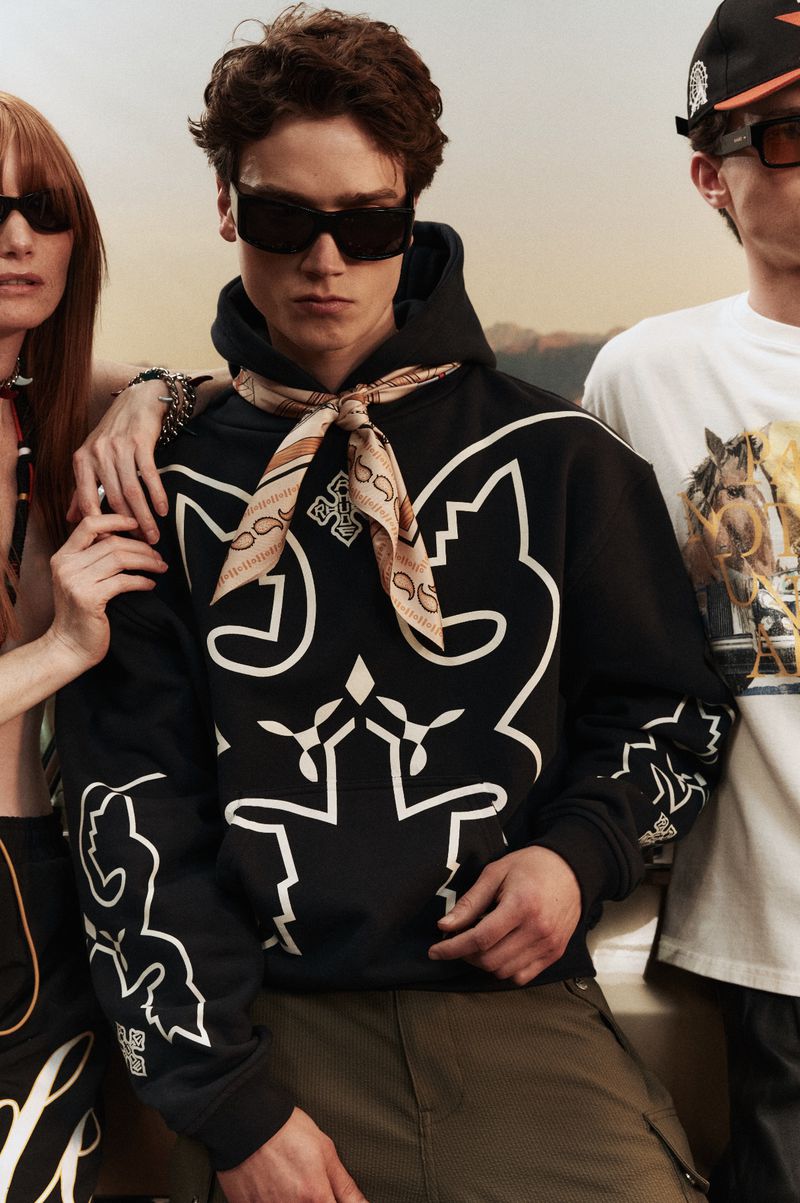 Three fashion models wearing sunglasses and patterned tops pose in front of a scenic background in a medium studio shot.