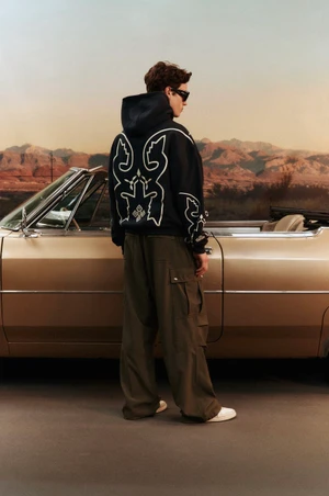 Man wearing a black hoodie and cargo pants standing beside a vintage convertible car against a desert landscape backdrop.