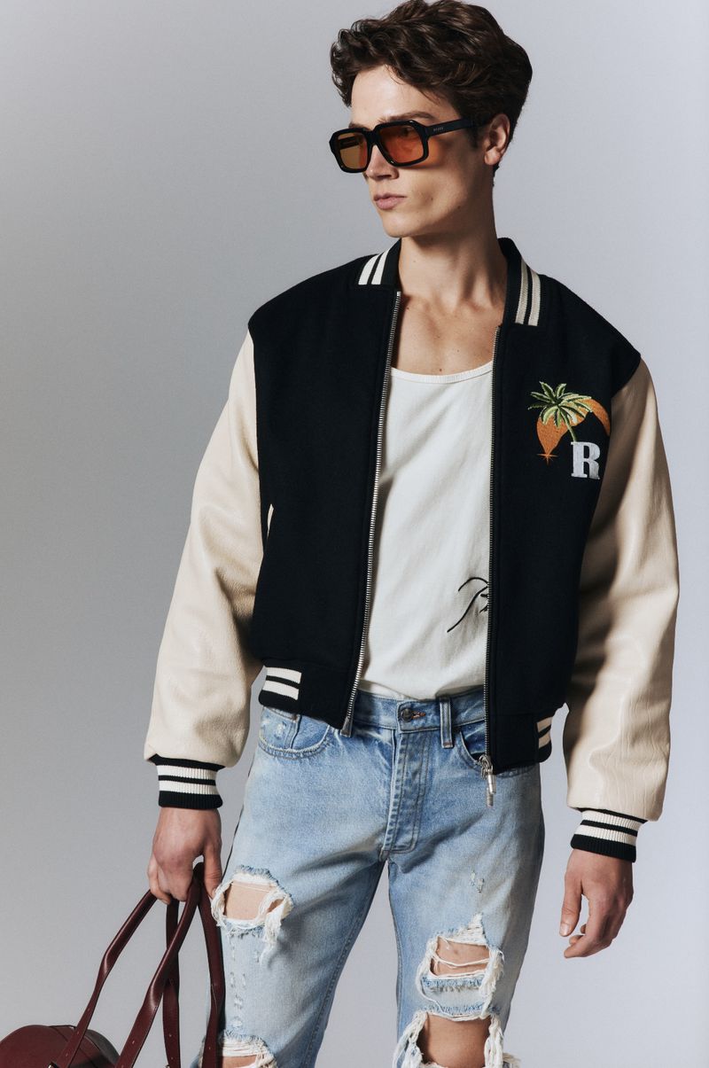 A young adult male poses in a black and beige varsity jacket, ripped jeans, and sunglasses, carrying a tote bag against a grey background.