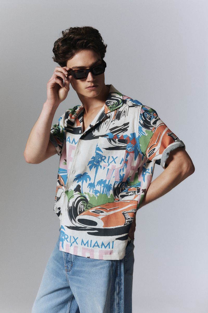 Man poses in short-sleeve button-down shirt with race car pattern, light jeans, sunglasses, and a neutral backdrop in a fashion editorial.