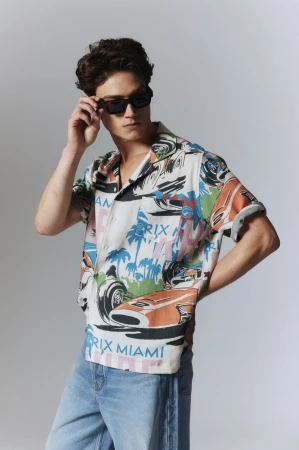 Man poses in short-sleeve button-down shirt with race car pattern, light jeans, sunglasses, and a neutral backdrop in a fashion editorial.