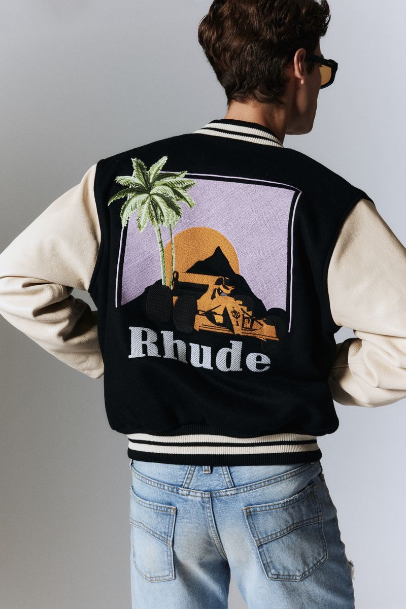 Model showcases a black bomber jacket with a tropical sunset graphic. Light blue jeans and a gray background complement the fashion shot.