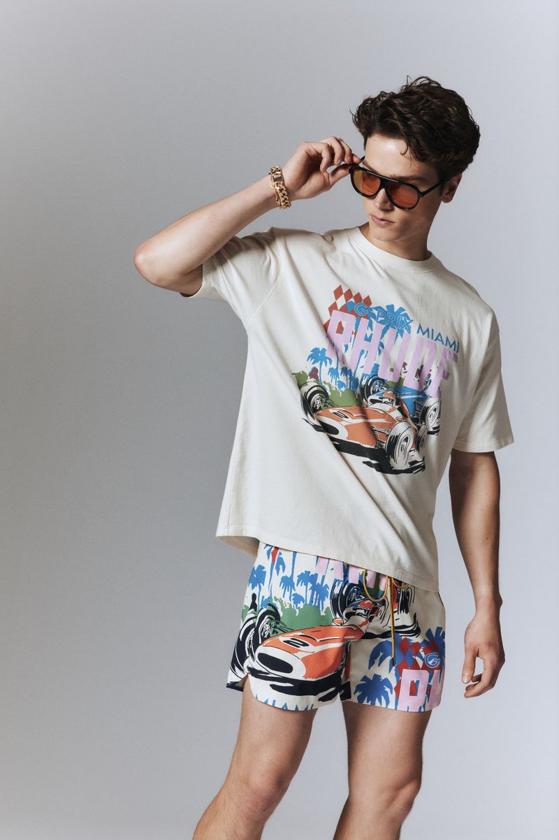 Man in a t-shirt and swim trunks with a race car print, accessorized with sunglasses and a gold bracelet, poses in a studio.