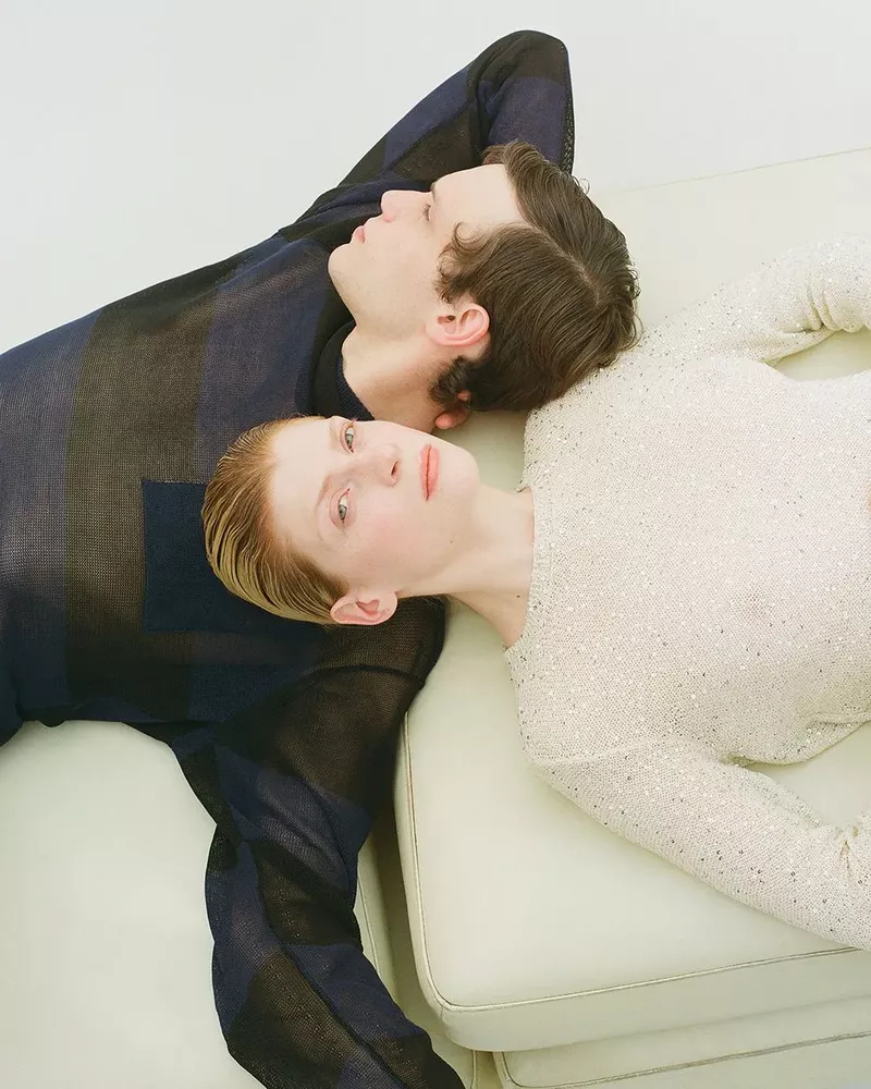 Two fair-skinned adults lie on a cream sofa in a studio. One wears a sheer blue striped top; the other wears a sparkly white long-sleeve top.