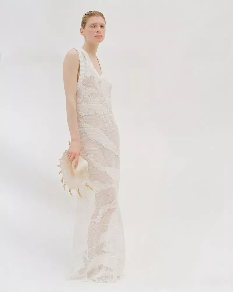 Elegant woman in sheer white dress holds large seashell in studio shot, creating a minimalist and ethereal fashion aesthetic.