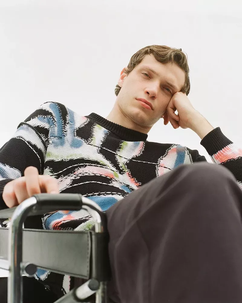 Portrait of a young adult man with curly hair, wearing a patterned sweater, and sitting in a wheelchair against a plain white background.