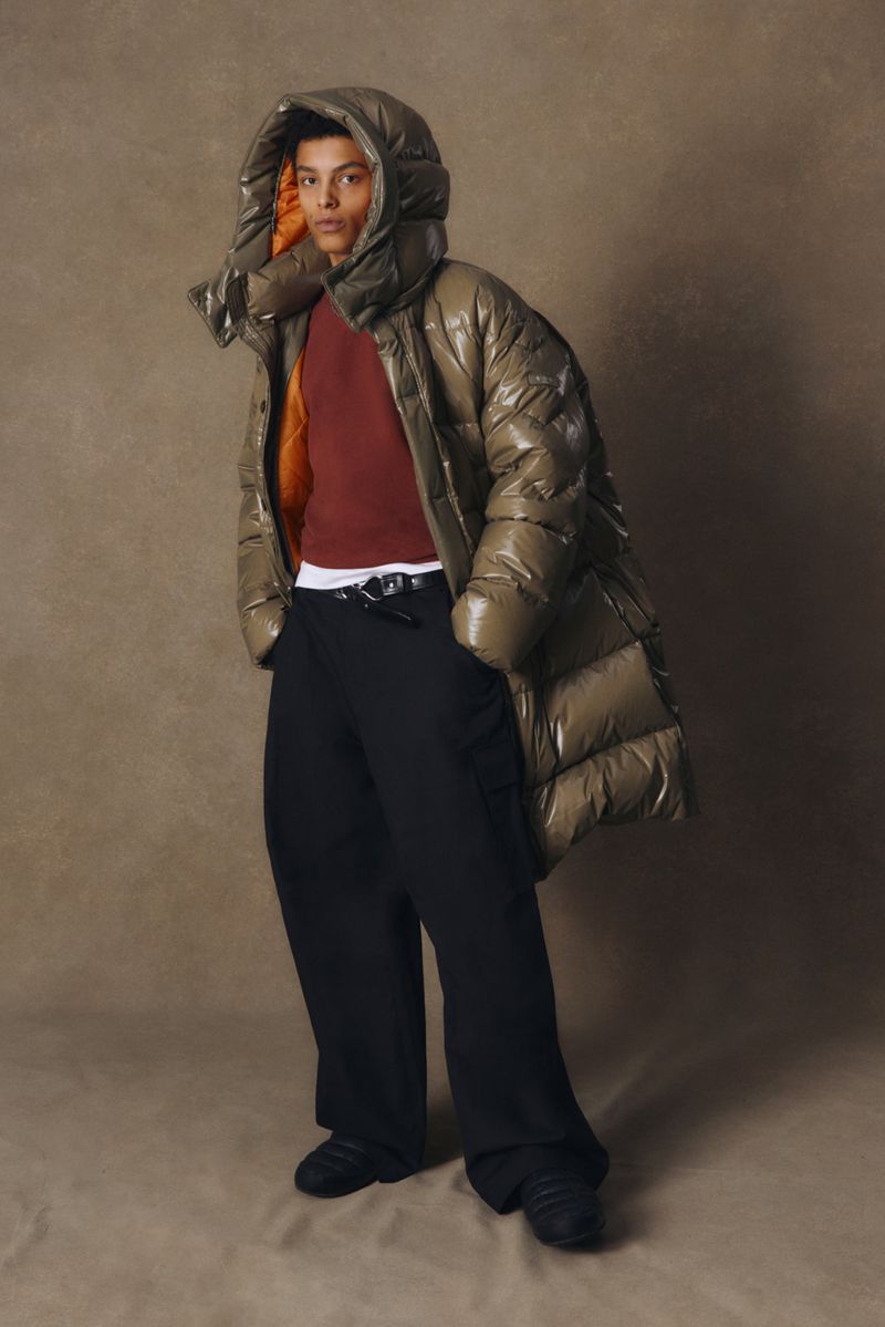 An adult stands indoors against a neutral backdrop, wearing a long, puffy, hooded olive-green coat with black pants and sneakers.