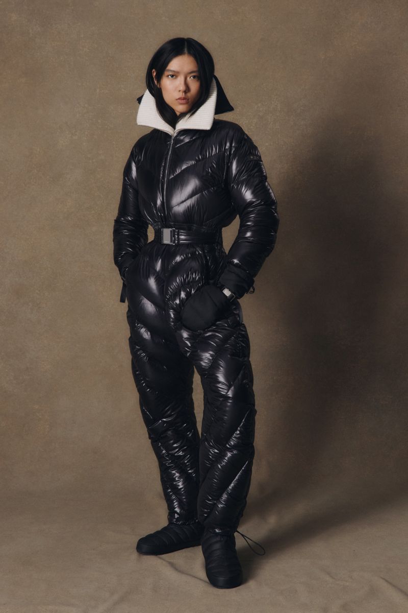 A fashionable Asian woman models a shiny black, quilted, belted snowsuit with a white collar in a neutral studio setting.