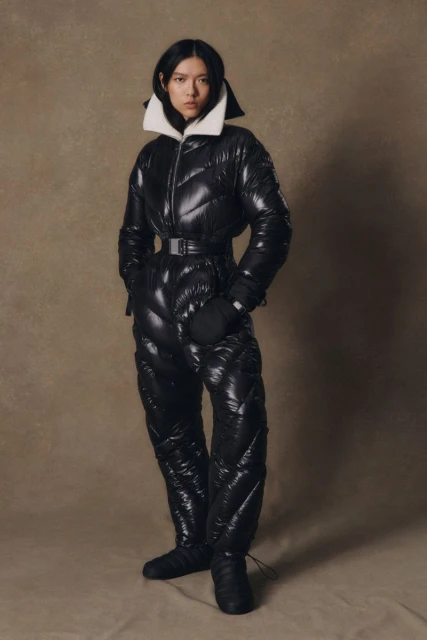 A fashionable Asian woman models a shiny black, quilted, belted snowsuit with a white collar in a neutral studio setting.